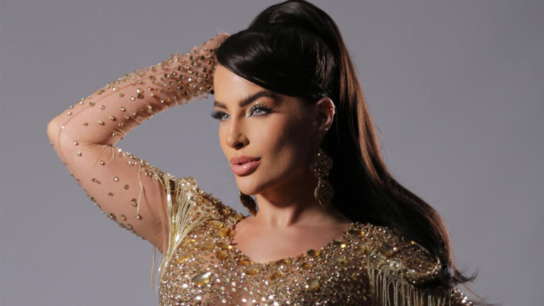 Mari Marquini com rabo de cavalo alto, usando vestido dourado com franjas e brilhos, posando com a mão na cabeça