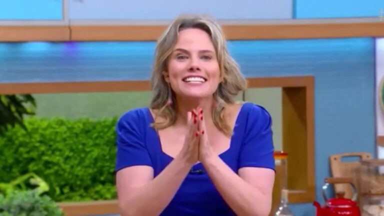 Mulher loira com cabelos na altura dos ombros sorri e junta as mãos enquanto apresenta programa de televisão em cenário colorido com tons de azul, verde e madeira ao fundo