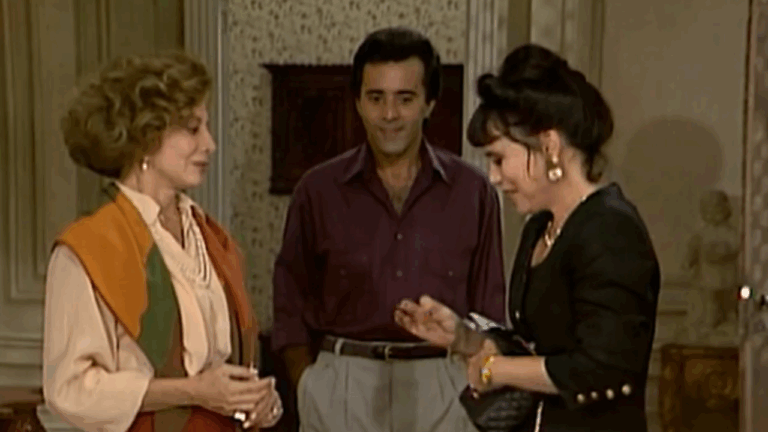 Cena mostra duas mulheres e um homem em uma sala decorada; as mulheres conversam cordialmente, enquanto o homem observa ao fundo sorrindo, em ambiente com tons clássicos e elegantes