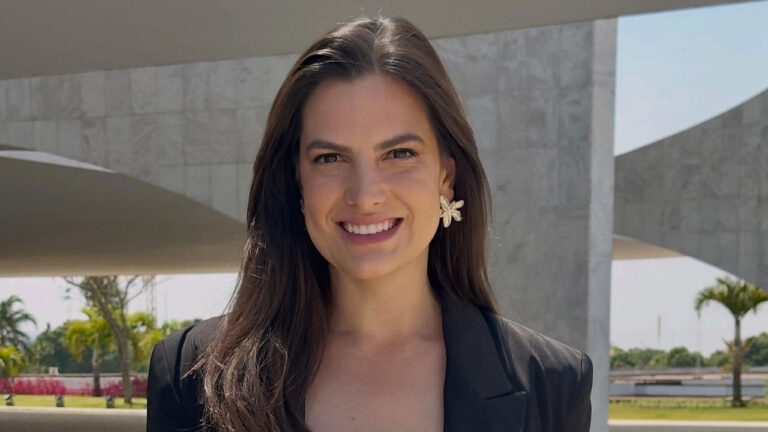 Jornalista Marina Demori sorrindo, de cabelo solto e blazer preto, usando brincos de flor, em área externa com construção de mármore