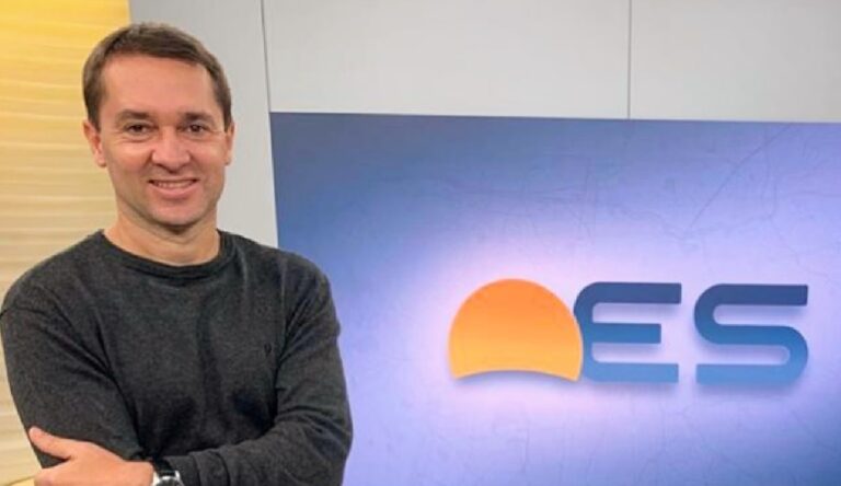 A imagem mostra um homem sorridente de braços cruzados, vestindo uma blusa escura, em um estúdio de televisão. Atrás dele, há um painel com o logotipo “ES”, que parece fazer parte de um programa jornalístico. O ambiente é bem iluminado e transmite uma atmosfera profissional