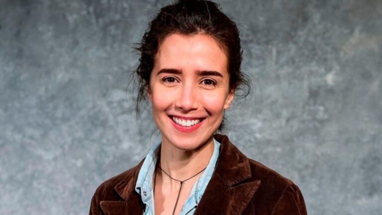 Mulher sorridente posa diante de fundo neutro, vestindo jaqueta marrom e camisa jeans clara