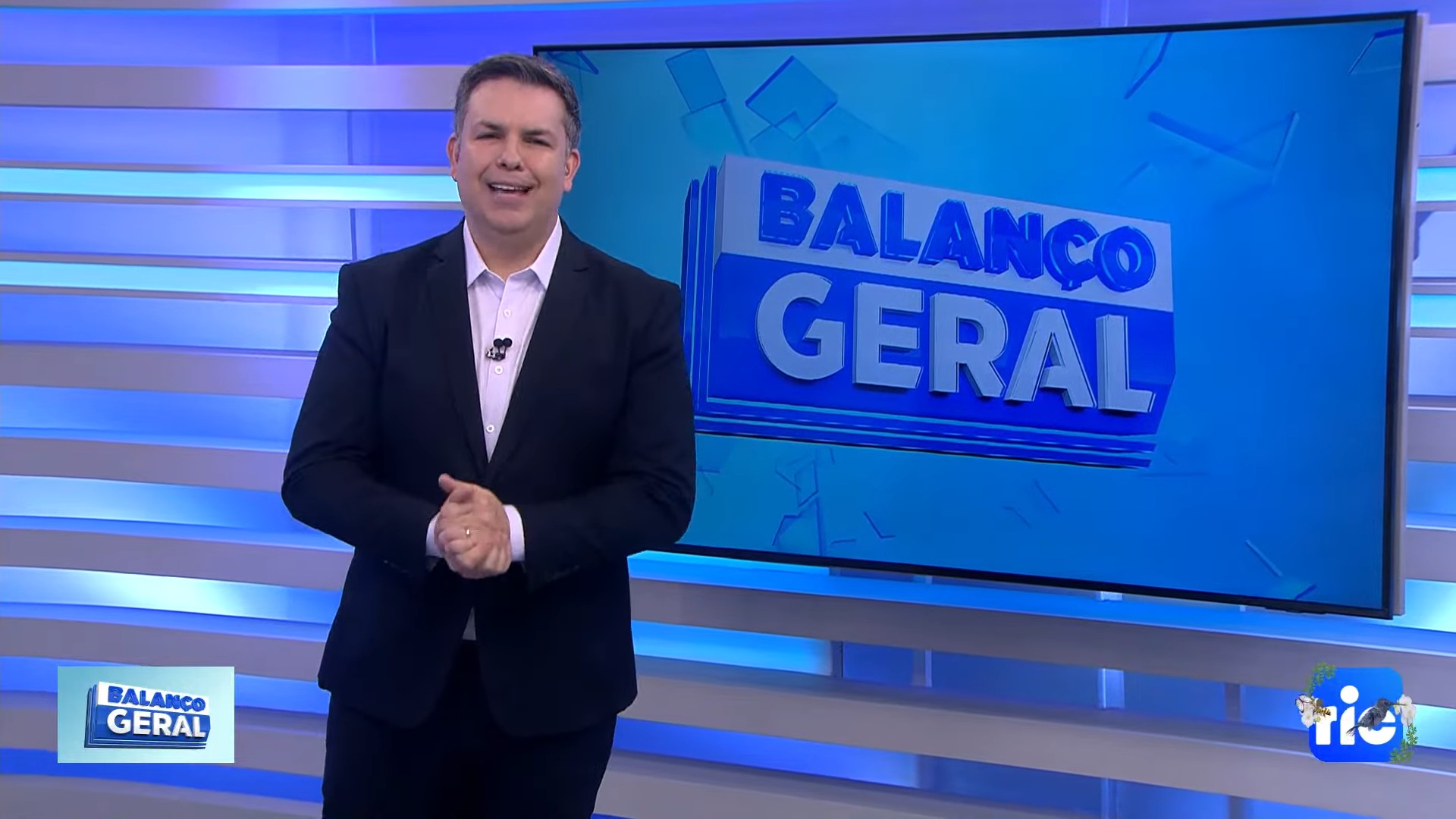 A imagem mostra Matheus Furlan no estúdio do programa Balanço Geral, diante de um grande painel azul com o logotipo do telejornal. Ele está vestindo terno escuro e camisa clara, sorrindo enquanto faz um gesto com as mãos, transmitindo uma postura confiante e acolhedora típica de um âncora de noticiário popular