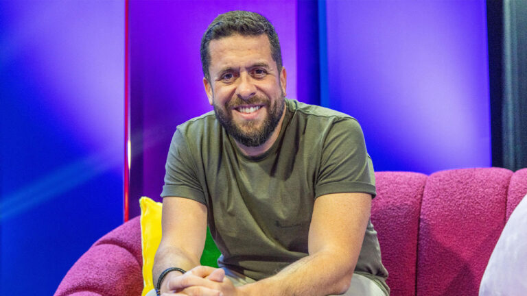 Maurício Meirelles sorrindo, de barba, sentado em um sofá rosa texturizado com fundo roxo neon no cenário do Aberto ao Público da Globo