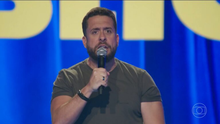 Maurício Meirelles, que é um homem de barba, segura microfone enquanto fala em um palco iluminado do programa Aberto ao Público, com luz azul e amarela, usando camiseta verde-oliva; no canto, aparece o logotipo da TV Globo