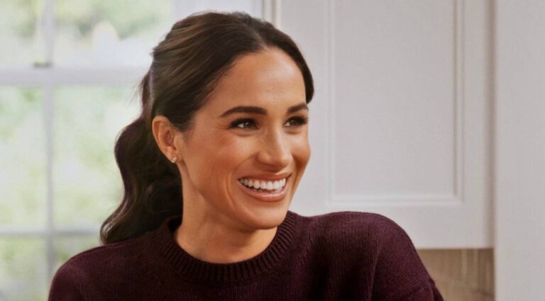 Meghan Markle sorridente com cabelo preso e blusa vinho aparece em ambiente interno iluminado por luz natural, com parede clara e janela ao fundo