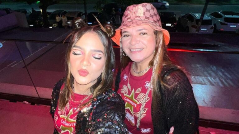 Duas mulheres posam para uma selfie em ambiente noturno iluminado por luzes coloridas. Ambas vestem camisetas vermelhas de camarote com paetês ou brilho por cima. A mulher à esquerda faz biquinho com os olhos fechados e usa penteado com dois coques altos. A mulher à direita sorri para a câmera e usa um chapéu estampado. Ao fundo, aparecem carros estacionados e a rua desfocada