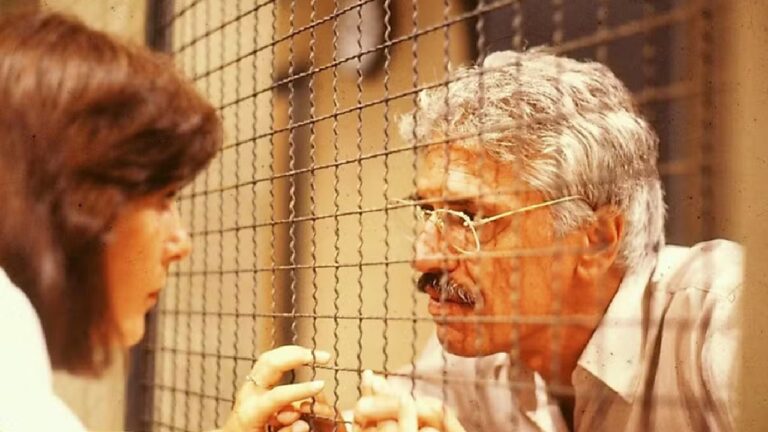 A imagem mostra uma cena intensa entre um homem e uma mulher separados por uma grade de arame. O homem, de cabelos grisalhos e usando óculos, está do lado de dentro, aparentando estar preso, enquanto a mulher o encara com expressão preocupada