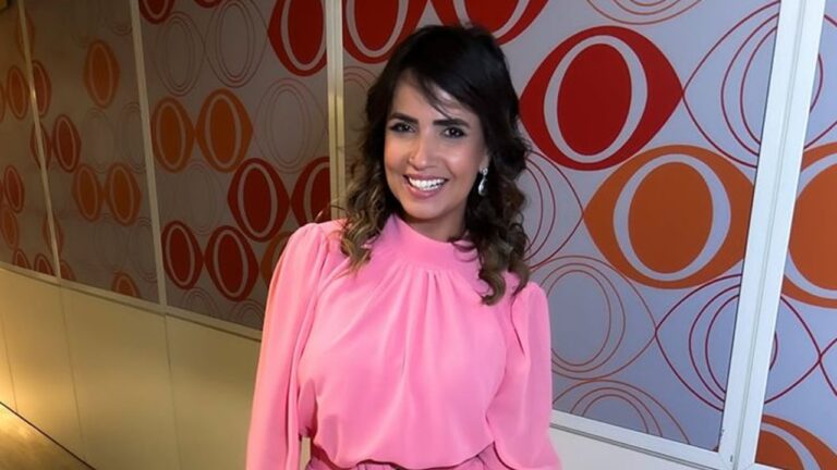 A imagem mostra uma mulher sorridente em um corredor decorado com painéis que têm círculos e formas ovais nas cores laranja e vermelho. Ela está vestindo um vestido rosa de mangas longas e cabelo solto com ondas. O ambiente parece ser interno, bem iluminado e com um ar moderno