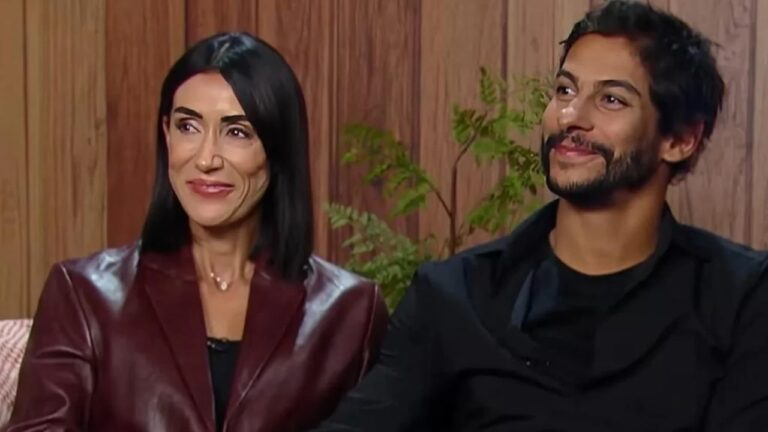A imagem mostra duas pessoas sentadas lado a lado, sorrindo e trocando um olhar simpático. A mulher, de cabelo preto e liso, veste um blazer de couro vinho, enquanto o homem, de barba e cabelo curto, usa uma camisa preta