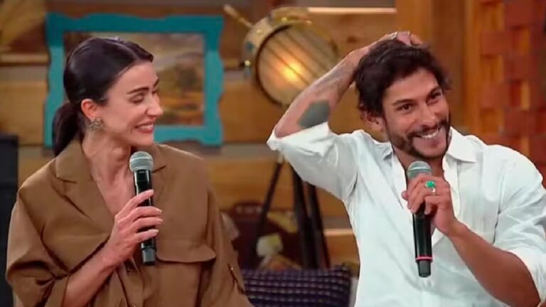 Michelle Barros e Shia sorriem enquanto seguram microfones em um programa de TV, sentados lado a lado em um cenário com decoração rústica e luz quente