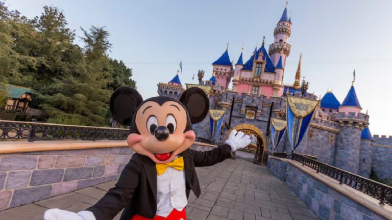 A imagem mostra o personagem Mickey Mouse posando sorridente em frente ao Castelo da Bela Adormecida, no parque Disneyland. Ele usa seu traje clássico com calça vermelha, casaco preto, colete branco e gravata borboleta amarela