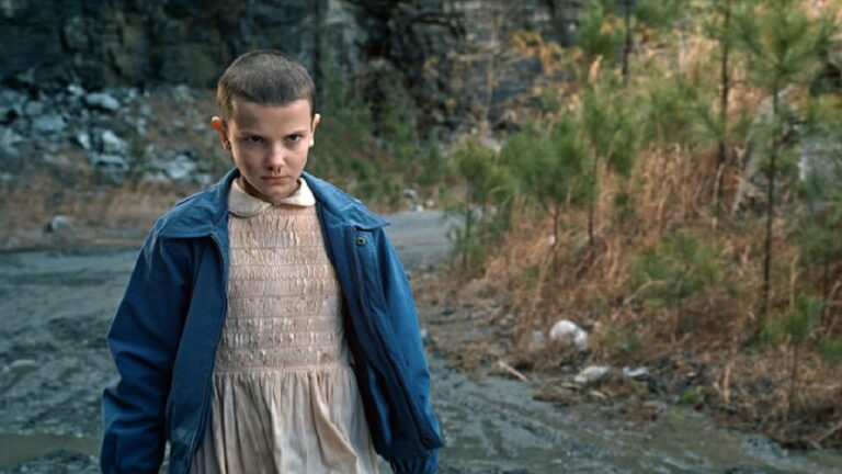 Criança de cabeça raspada aparece com expressão séria, vestindo vestido rosa claro e jaqueta azul, em uma estrada cercada por árvores e rochas, em cena de atmosfera tensa