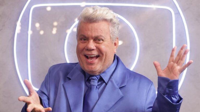 Carnavalesco Milton Cunha sorridente, com cabelo branco e terno azul-claro, de braços abertos diante do logo neon da Globo