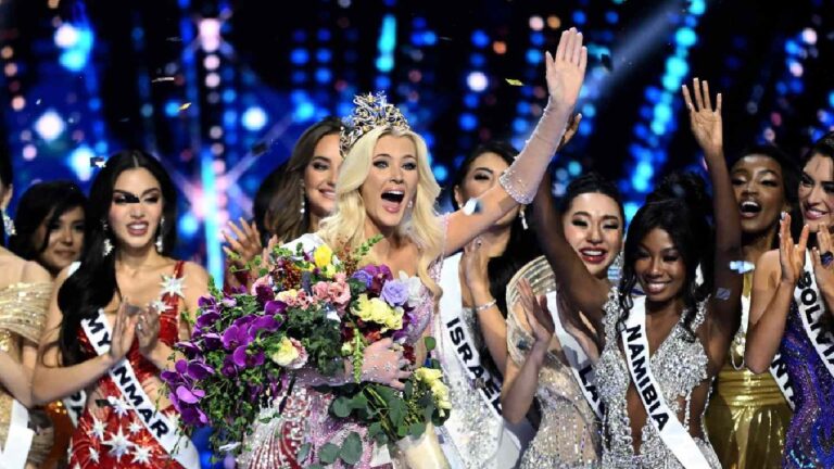 Mulher coroada comemora sorridente com um buquê de flores coloridas nos braços, enquanto outras candidatas a concursos de beleza aplaudem ao redor, usando faixas com nomes de países. O cenário é iluminado por luzes azuladas e confetes, representando o momento de coroação em um concurso internacional