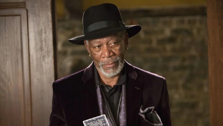 A imagem mostra um homem mais velho usando chapéu preto, terno escuro e camisa igualmente escura, segurando um baralho de cartas