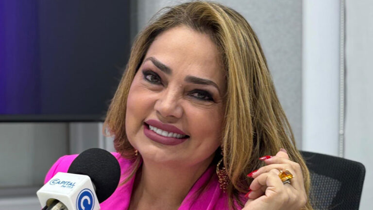 Apresentadora Nani Venâncio sorrindo com blazer pink, maquiagem carregada e cabelo loiro em estúdio da rádio Capital FM