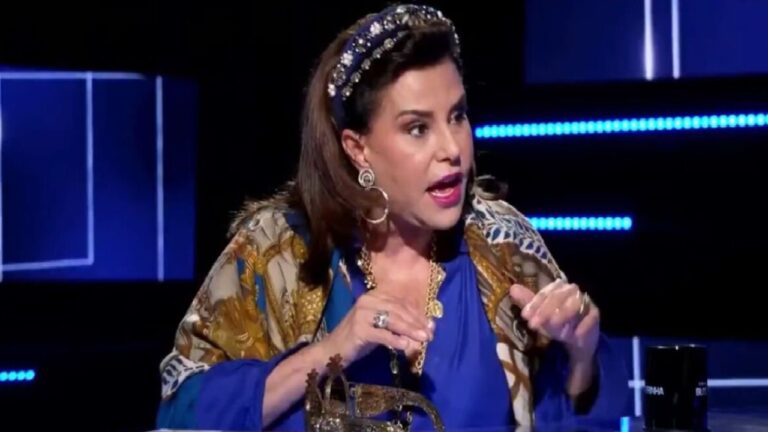 Narcisa Tamborindeguy vestida de azul e com tiara brilhante fala com expressão enfática em um estúdio iluminado, diante de uma mesa com adereço dourado e caneca preta