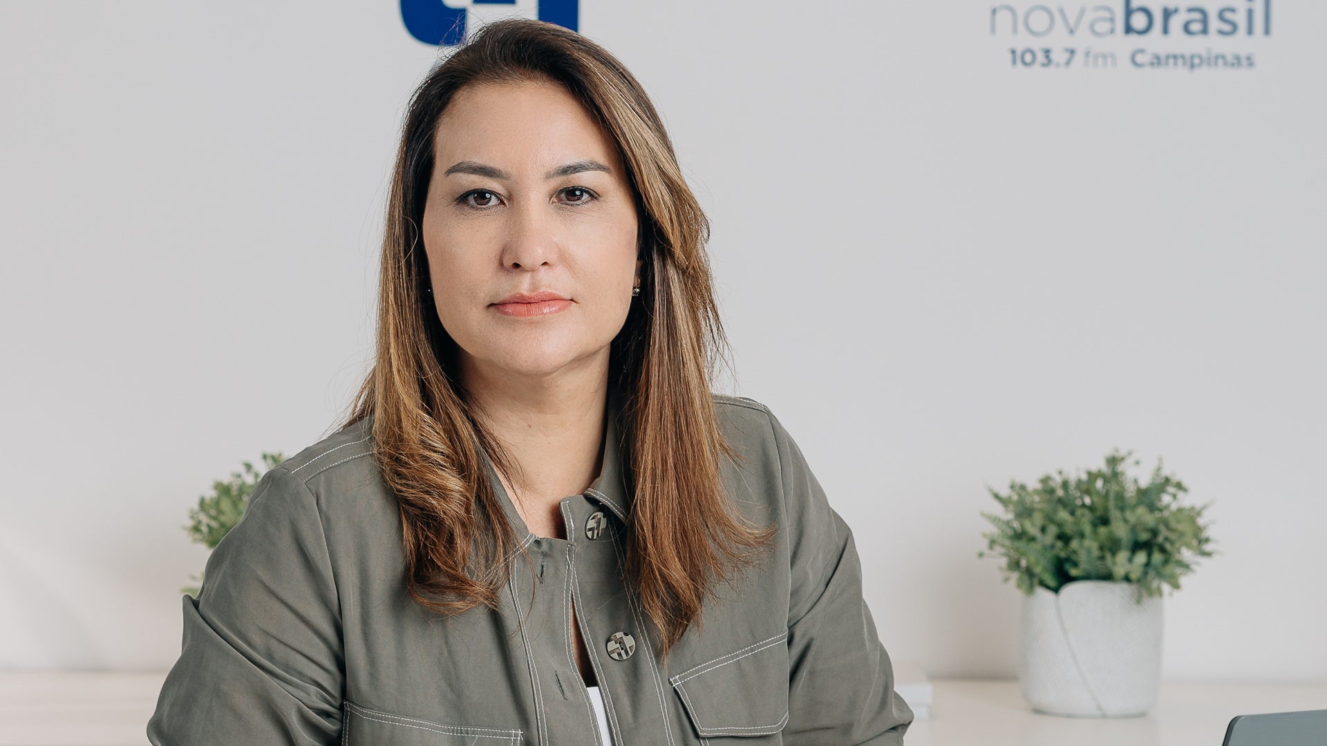 A imagem mostra Nathalie Pilan Caram, uma mulher de expressão neutra, com cabelos castanhos claros e lisos, usando uma camisa verde-acinzentada. Ela está sentada em um ambiente corporativo claro, com vasos de plantas ao fundo e o logotipo da NovaBrasil 103.7 FM Campinas visível na parede. A foto tem iluminação suave e transmite um tom profissional e sereno