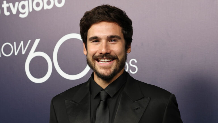 Ator Nicolas Prattes com terno preto e gravata sorrindo em evento da TV Globo para celebrar os 60 anos da emissora