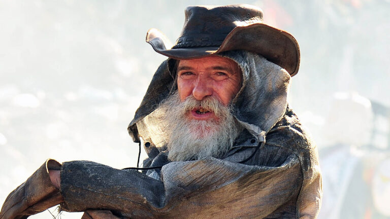 Ator José de Abreu como Nilo com barba grisalha longa e chapéu de couro, em cena do lixão da novela Avenida Brasil
