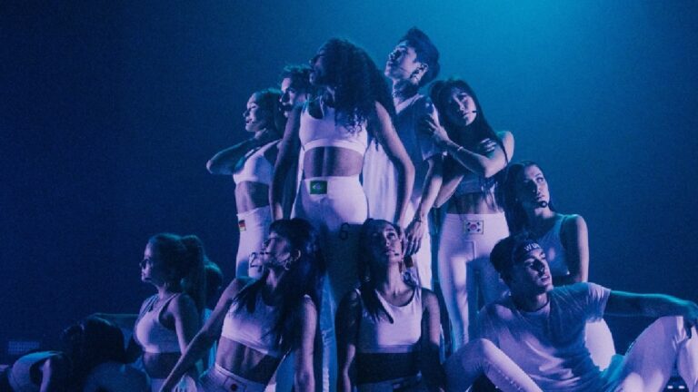 Grupo de dançarinos no palco faz pose coreografada sob iluminação azul; todos vestem roupas brancas esportivas com bandeiras de diferentes países, transmitindo energia e unidade em apresentação artística