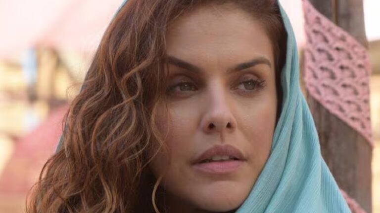 Mulher de expressão séria usa um véu azul claro sobre os cabelos castanhos, em ambiente externo ensolarado. A cena transmite um clima contemplativo e remete a produções históricas ou de época ambientadas em regiões desérticas