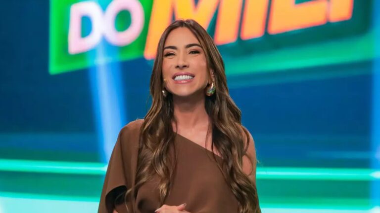 Patricia Abravanel apresenta programa de TV em estúdio colorido, com iluminação verde e azul ao fundo e parte de letreiro visível atrás dela