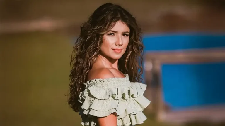 Mulher jovem com cabelos longos e cacheados posa ao ar livre sob luz dourada. Ela usa uma blusa verde-clara ombro a ombro com babados e olha para a câmera com expressão serena. Ao fundo, aparece um cenário desfocado com gramado e uma estrutura azul