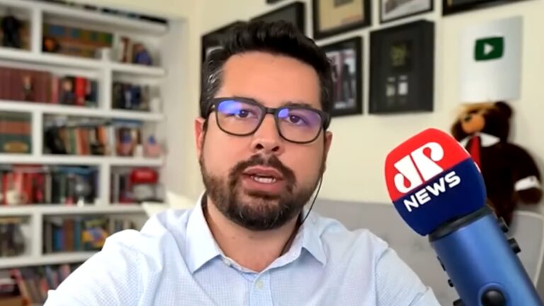 A imagem mostra Paulo Figueiredo, um homem usando óculos e camisa social clara, falando em frente a um microfone com o logotipo da Jovem Pan News. Ao fundo, há uma estante com livros e quadros decorativos, sugerindo um ambiente de home office