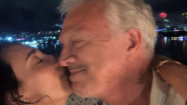 A imagem mostra um casal trocando um beijo carinhoso no rosto durante a noite. A mulher, de olhos fechados, abraça o homem, que sorri com expressão de ternura. Ao fundo, é possível ver luzes coloridas refletindo na água, sugerindo um cenário urbano iluminado, possivelmente próximo a um rio ou à orla de uma cidade