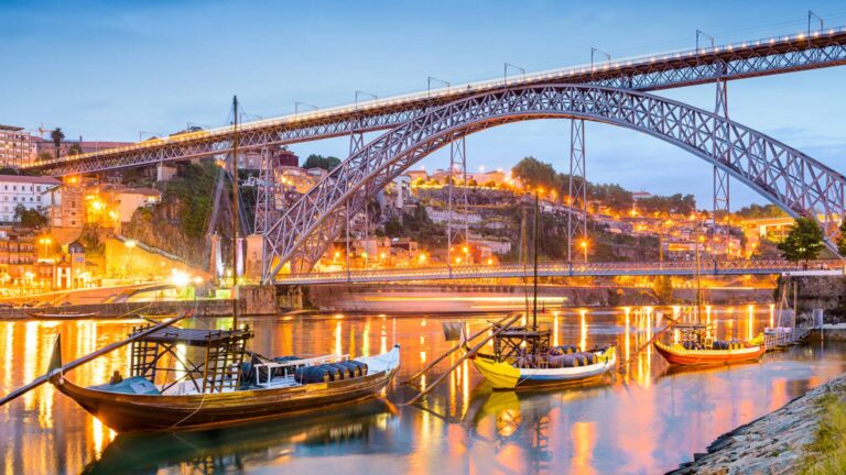 Essa imagem mostra a Ponte Dom Luís I, um dos marcos mais icônicos da cidade do Porto, em Portugal. A ponte cruza o rio Douro e conecta o Porto a Vila Nova de Gaia. Na cena, vê-se o reflexo das luzes da cidade sobre o rio, barcos tradicionais (chamados rabelos) e uma bela iluminação ao entardecer, destacando a arquitetura metálica da ponte