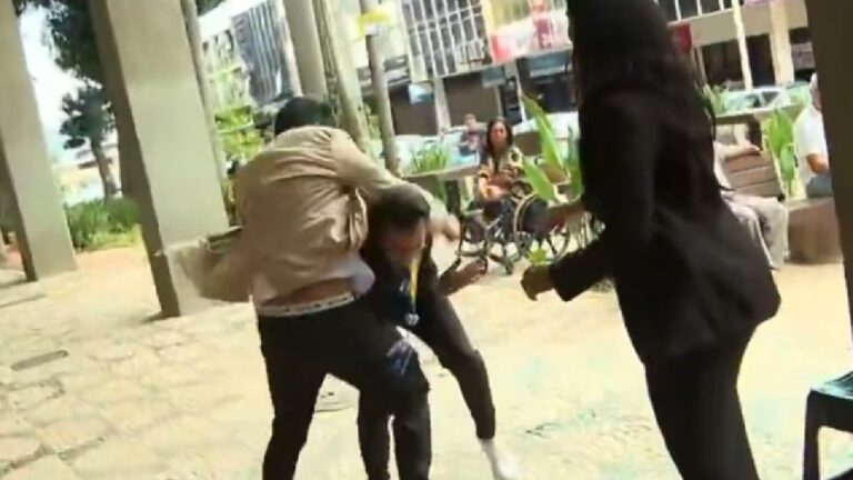 Imagem mostra uma confusão em via pública, com duas pessoas em luta corporal enquanto outra tenta intervir; ao fundo, pedestres observam a cena em uma área urbana com prédios e calçada coberta