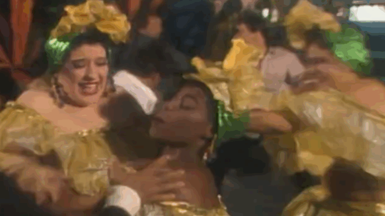 Mulheres com vestidos dourados e adereços de cabeça dançam em cena festiva de Rainha da Sucata