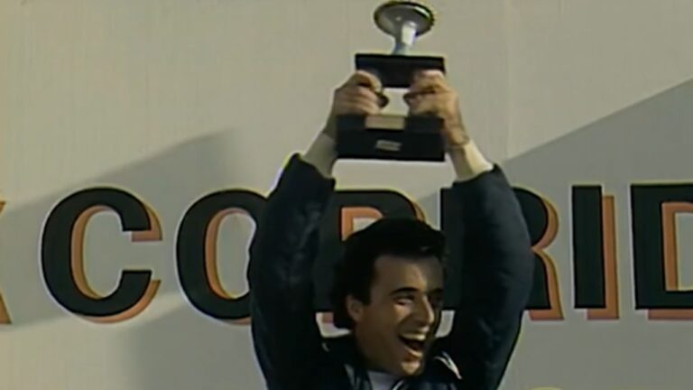 A imagem mostra um homem sorridente erguendo um troféu acima da cabeça em um pódio de premiação. Ele veste um macacão de corrida azul e parece comemorar uma vitória esportiva. Ao fundo, há parte de um painel com letras grandes que indicam um evento competitivo