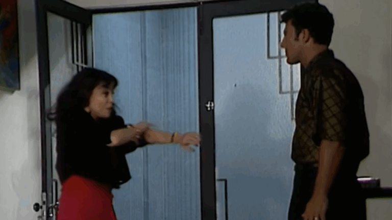 Mulher faz gesto enfático com os braços diante de um homem, em ambiente interno iluminado, próximo a uma porta de vidro; a cena sugere uma conversa intensa ou discussão