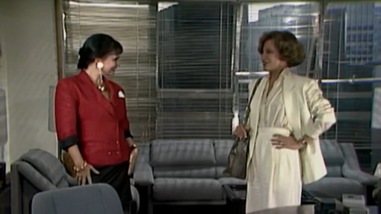 A imagem mostra duas mulheres conversando em um ambiente corporativo elegante, provavelmente um escritório. Uma delas veste um conjunto branco e carrega uma bolsa, enquanto a outra usa um blazer vermelho e brincos grandes dourados. Elas parecem estar em uma conversa amigável, com expressões de simpatia. O cenário ao fundo tem janelas amplas com persianas e móveis de escritório, sugerindo uma cena de novela ou série dos anos 1980 ou 1990