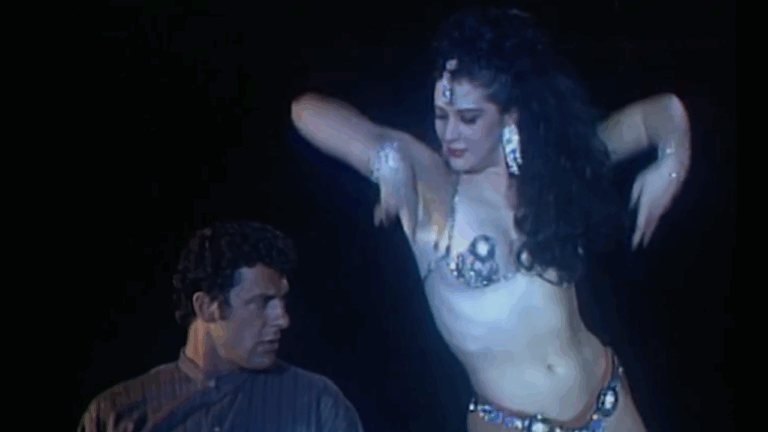 Cena mostra uma dançarina realizando uma dança sensual em frente a um homem que a observa, em ambiente escuro com iluminação teatral