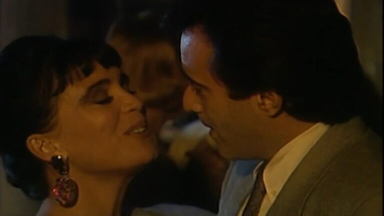 Mulher com brincos grandes sorri de olhos fechados enquanto conversa de perto com homem de terno em ambiente noturno