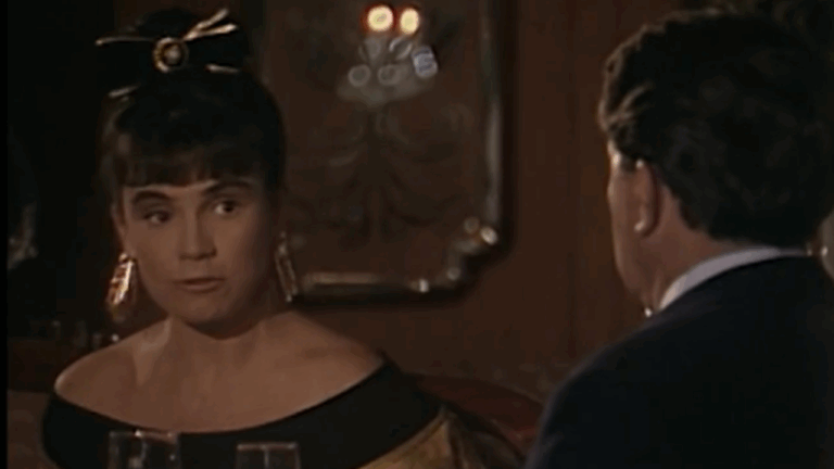 Cena mostra uma mulher e um homem sentados em uma mesa de restaurante elegante, com iluminação baixa e taças à frente. A mulher usa vestido preto de ombros à mostra, brincos longos e um laço no cabelo, enquanto fala com o homem à sua frente