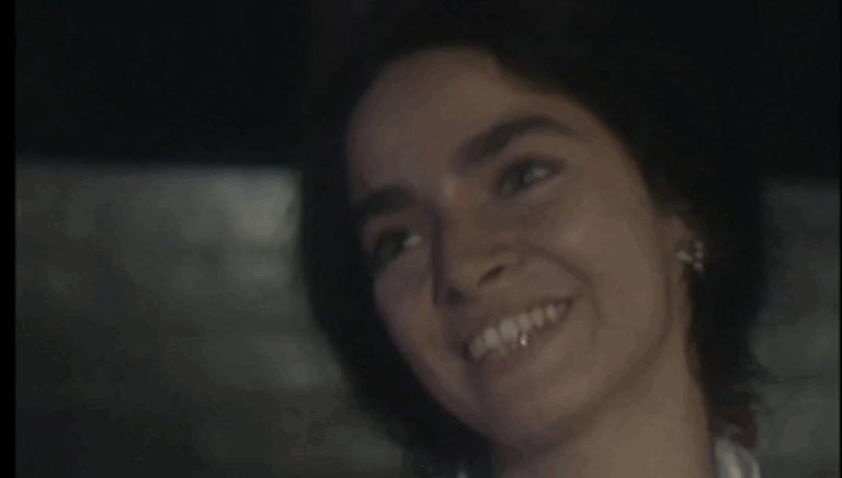 A imagem mostra uma mulher sorrindo levemente em um ambiente com pouca iluminação. Ela tem cabelos curtos e escuros, usa brincos delicados e parece olhar para alguém fora de quadro, transmitindo uma expressão de ternura e tranquilidade