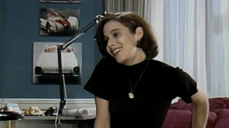 Mulher jovem, de cabelos curtos castanhos, sorri inclinando levemente a cabeça em um ambiente interno. Ela veste blusa preta e colar dourado. Ao fundo, há uma parede azul com quadros de carros esportivos, uma luminária articulada sobre a mesa e cortinas claras ao lado de um sofá vermelho.