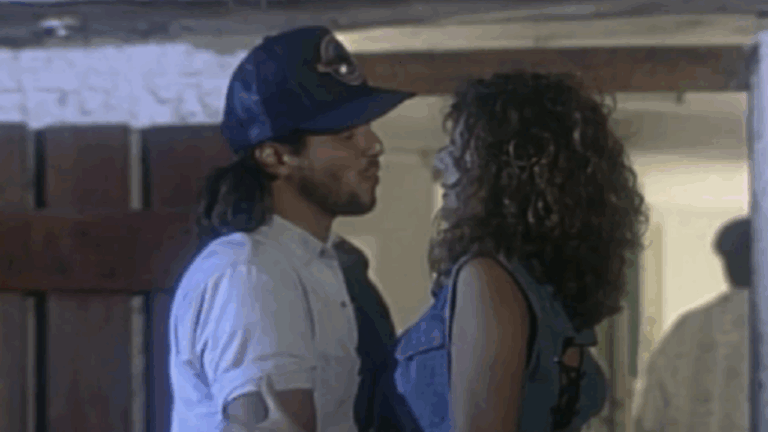 A imagem mostra um homem e uma mulher próximos um do outro, em um ambiente interno com iluminação suave. O homem usa boné azul e camisa clara, enquanto a mulher tem cabelos cacheados e veste uma blusa jeans sem mangas