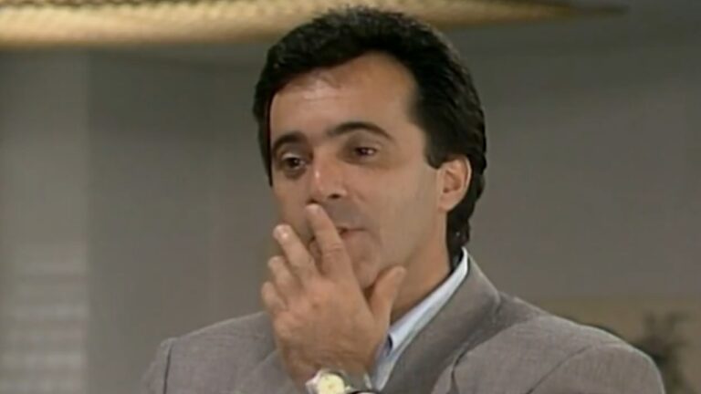 Homem de terno cinza leva a mão ao rosto com expressão pensativa em ambiente interno iluminado