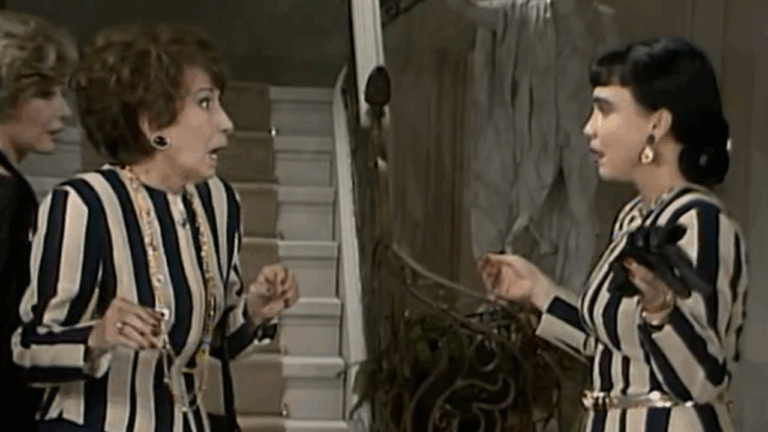 Duas mulheres em um ambiente interno discutem próximas a uma escada. Ambas usam roupas listradas em preto e branco. A mulher à esquerda demonstra surpresa, com expressão de espanto e mãos erguidas. A mulher à direita gesticula enquanto segura um objeto escuro. Ao fundo, vê-se a escadaria e parte da decoração da casa.