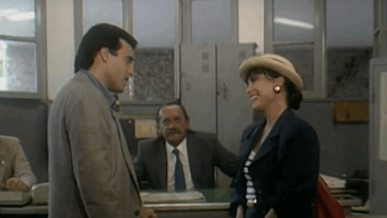 Cena mostra três pessoas em um escritório antigo: um homem e uma mulher em pé, sorrindo um para o outro, enquanto outro homem, sentado atrás de uma mesa, os observa. O ambiente tem janelas grandes, armários metálicos e iluminação típica de repartições públicas, sugerindo um clima leve e cotidiano