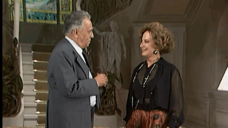 Cena mostra um homem e uma mulher mais velhos conversando em um ambiente elegante, com uma escadaria e estátua ao fundo. Ambos estão bem vestidos — ele de terno e ela com blusa preta translúcida e saia estampada —, trocando olhares amistosos em um clima de cordialidade