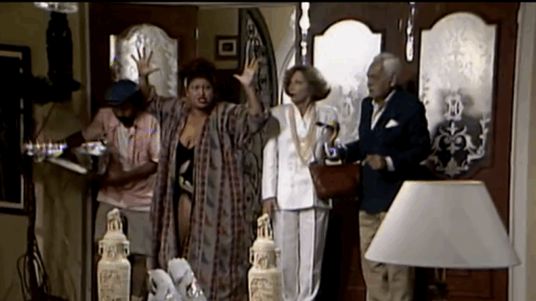 A imagem mostra uma cena de comédia em um ambiente doméstico, com quatro pessoas próximas à porta de entrada. Uma mulher ao centro parece muito exaltada, gesticulando com as mãos levantadas, enquanto os outros três observam com surpresa ou preocupação. O cenário tem decoração clássica, com vasos e móveis elegantes.