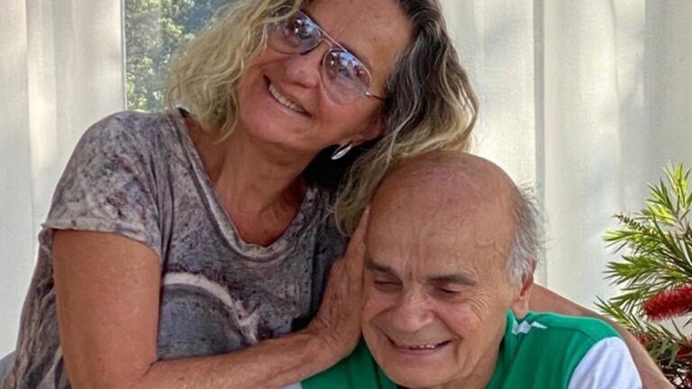 A imagem mostra uma mulher abraçando um homem sentado, ambos sorrindo de forma afetuosa. Eles estão em um ambiente iluminado, provavelmente ao ar livre, com cortinas claras e uma planta ao lado, transmitindo uma atmosfera de tranquilidade e carinho.