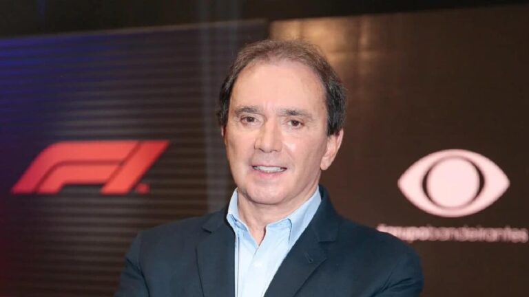 Homem de terno posa sorridente em frente a painéis com os logotipos da Fórmula 1 e de uma emissora de televisão, sugerindo evento ou cobertura esportiva